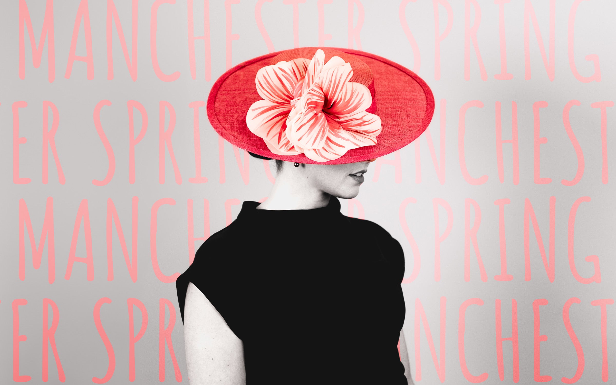 Model stands in front of a wall that reads Manchester Spring wearing a bright pink boater hat with silk poppy flowers with hand painted centers
